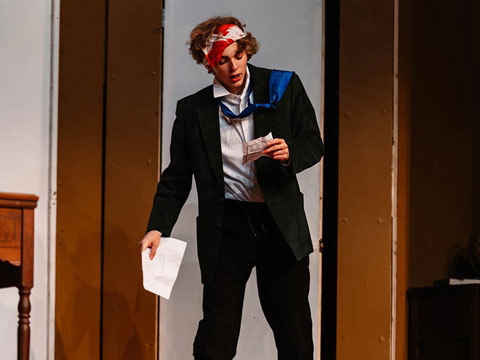 Person in a suit with a bandana, holding papers on stage.
