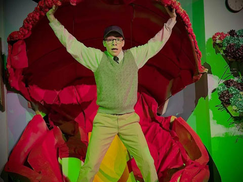 A person stands inside a large, red flower prop with arms raised, wearing a cap and glasses, against a green wall backdrop.