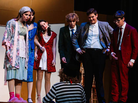 A group of five people in costumes stand on stage, looking at a person in a striped shirt seated in front of them.